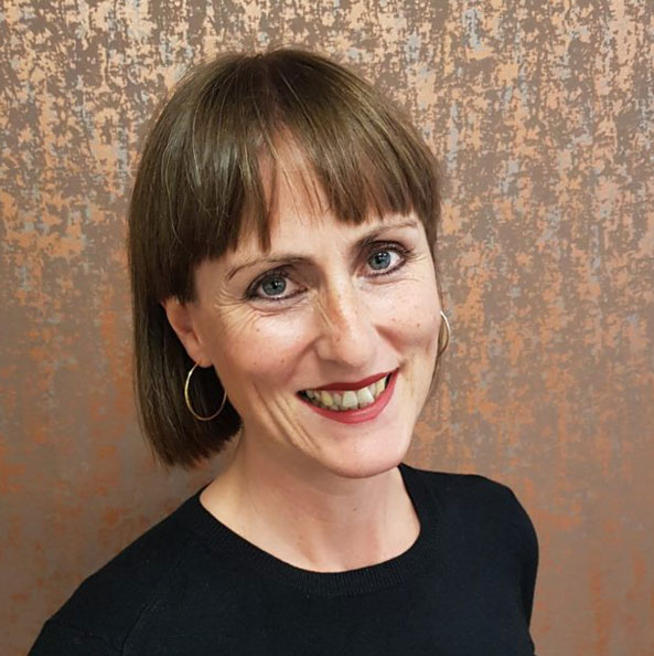 Photo of Head of Development Lou Benson. She is a white middle aged woman with short brown hair and a fringe. She is wearing a black tshirt and hooped earring and smiling to camera.