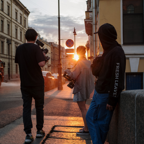 Three behind the scenes workers and standing by the side of an inner-city road whilst they watch and film the sunset in the distance.