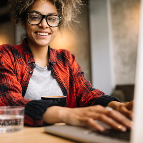 A woman is sat to a desk with a laptop and a mug. She is looking directly at the camera and is smiling.